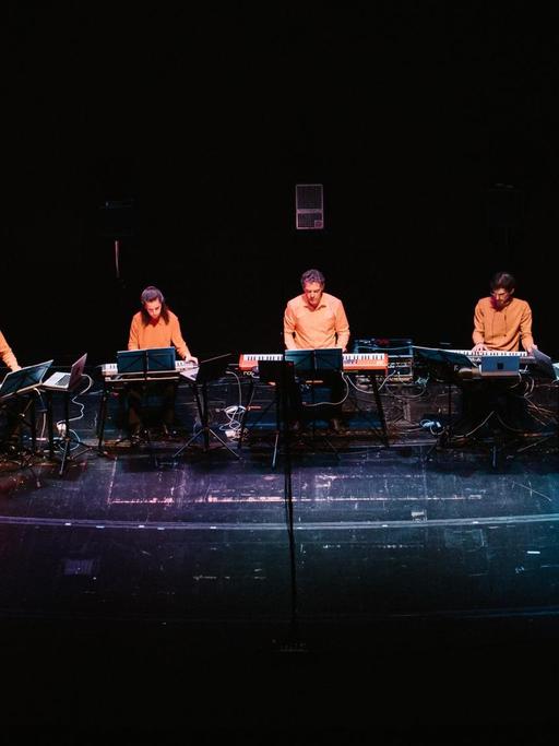 12 Musiker in orangefarbenen Hemden sitzen im Halbkreis auf der Bühne