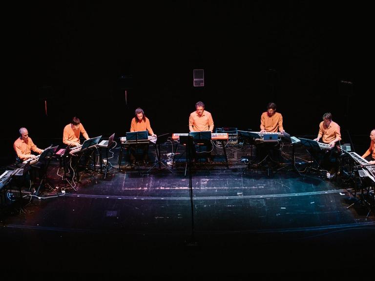 12 Musiker in orangefarbenen Hemden sitzen im Halbkreis auf der Bühne