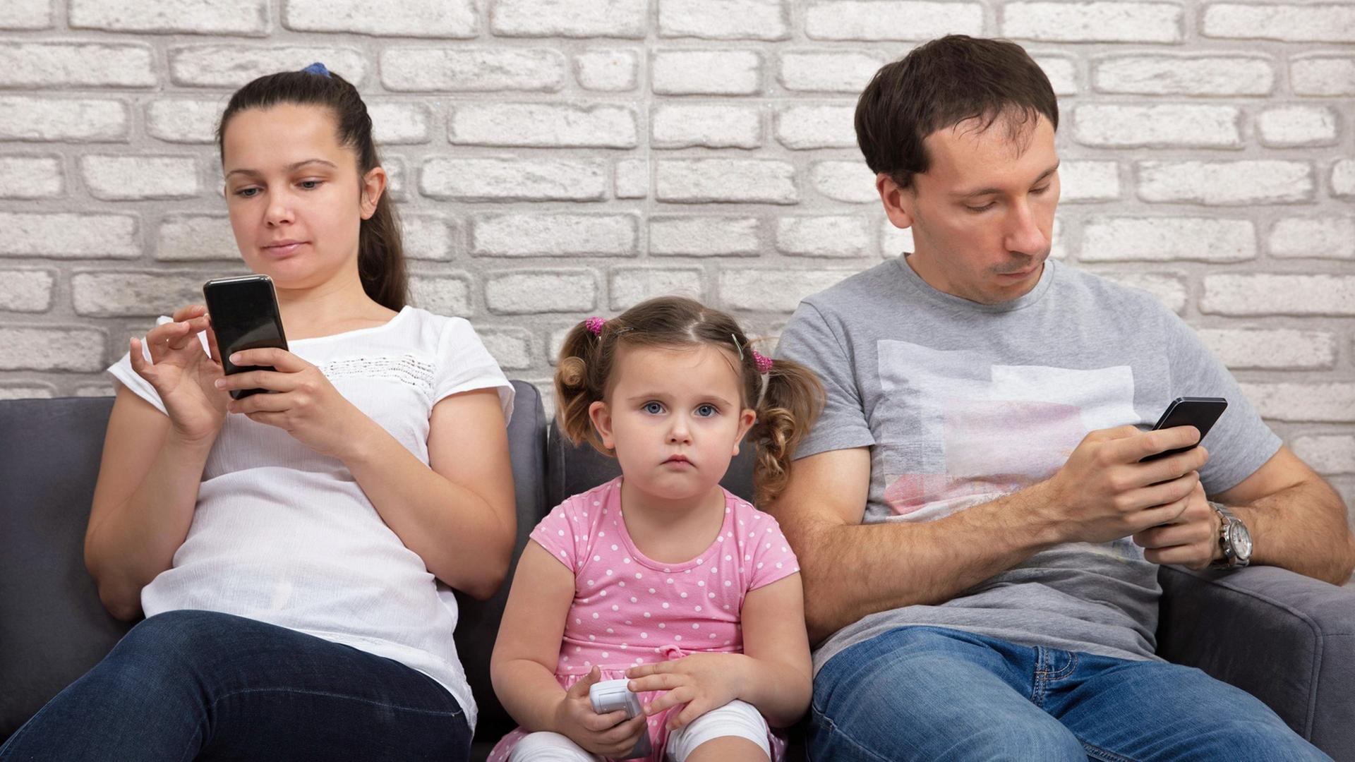 Ein Paar sitzt auf dem Sofa, beide sind mit ihrem Smartphone beschäftigt. Dazwischen sitzt ein kleines Mädchen. Ein Paar sitzt auf dem Sofa, beide sind mit ihrem Smartphone beschäftigt. Dazwischen sitzt ein kleines Mädchen.