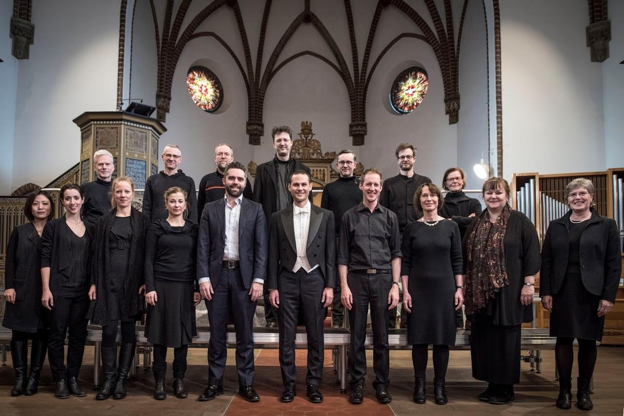Ein Chor steht mit Gast und Dirigenten in einem Kirchenraum. Ein Chor steht mit Gast und Dirigenten in einem Kirchenraum.