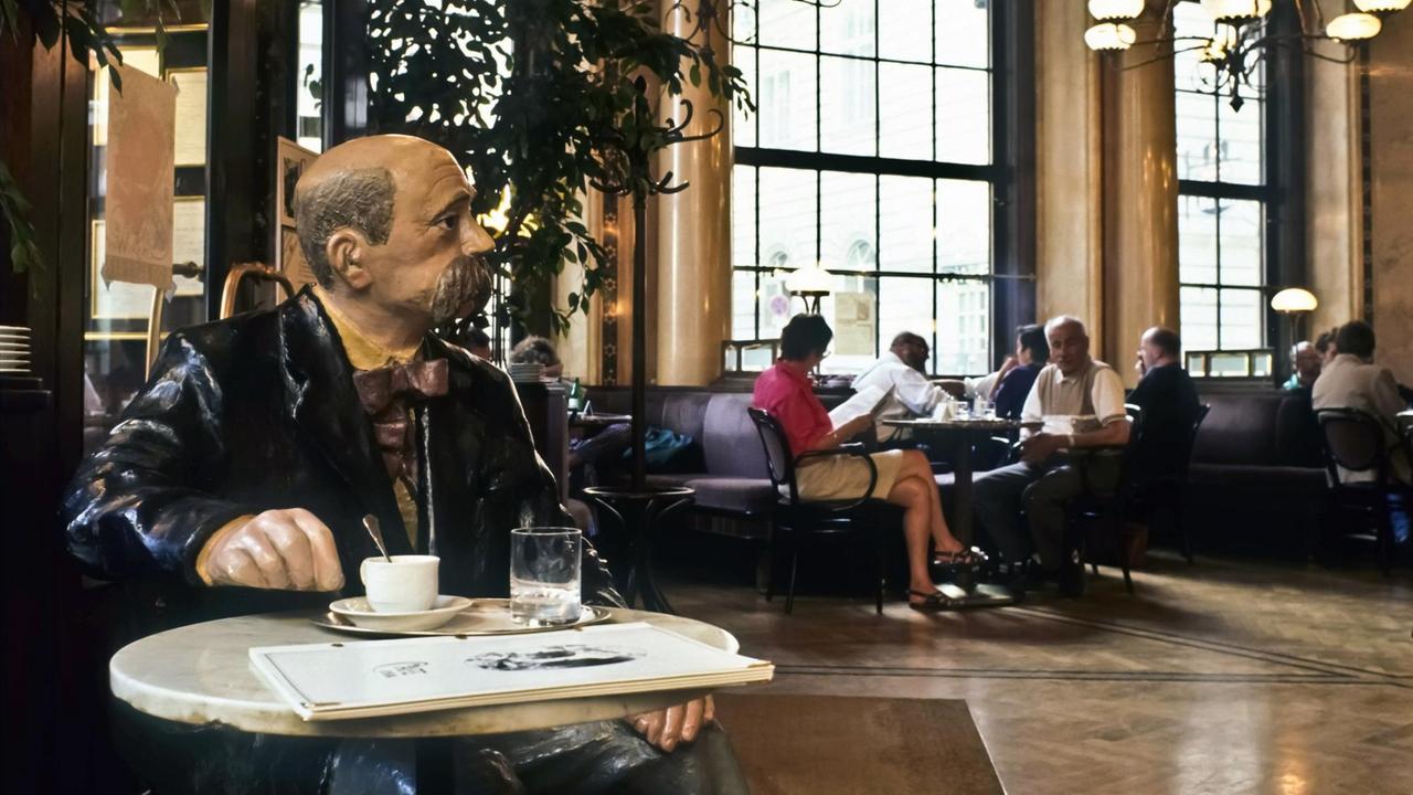 Eine Figur des österreichischen Schriftstellers Peter Altenberg sitzt mit einer Zeitung und einer Kaffeetasse an einem kleinen, runden Tisch im Wiener Café Central.