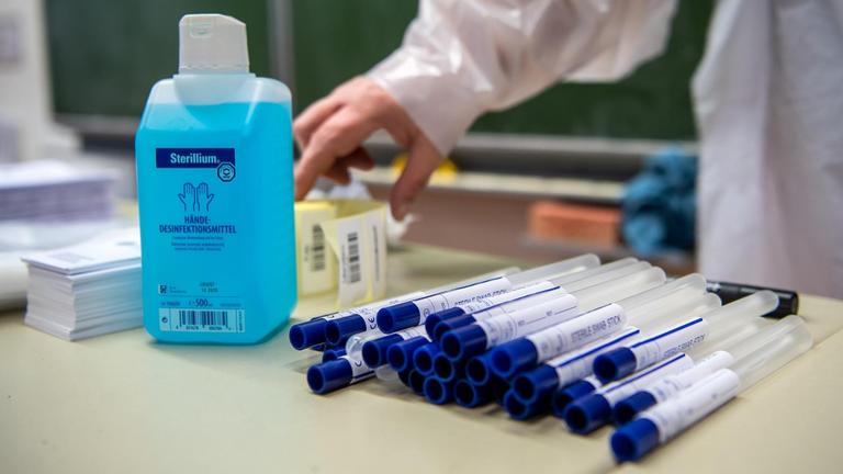 Eine Person mit Schutzhandschuhen und Kittel steht vor einem Tisch mit Teströhrchen und Desinfektionsmittel. Eine Person mit Schutzhandschuhen und Kittel steht vor einem Tisch mit Teströhrchen und Desinfektionsmittel.