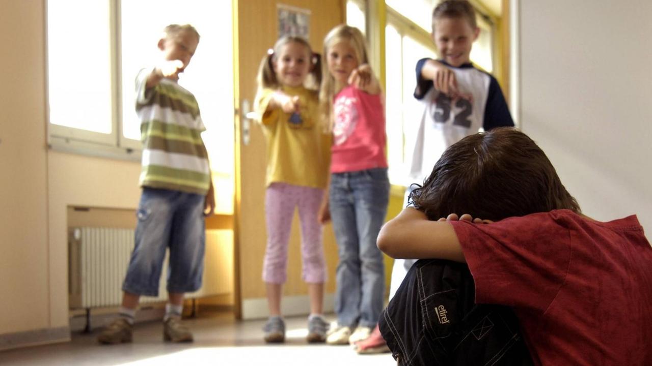 Ein Junge hockt traurig auf dem Boden eines Schulflurs, im Hintergrund zeigen Kinder mit dem Finger auf ihn und lachen Ein Junge hockt traurig auf dem Boden eines Schulflurs, im Hintergrund zeigen Kinder mit dem Finger auf ihn und lachen
