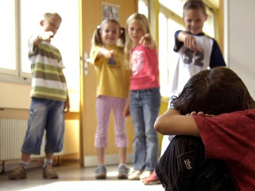 Ein Junge hockt traurig auf dem Boden eines Schulflurs, im Hintergrund zeigen Kinder mit dem Finger auf ihn und lachen