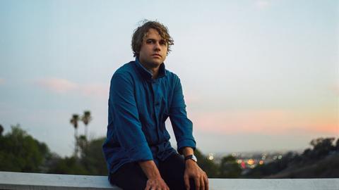 Der Musiker Kevin Morby sitzt auf einem Balken vor einem Tal, im Hintergrund leuchten Lichter einer Stadt.