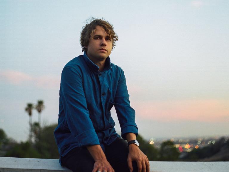Der Musiker Kevin Morby sitzt auf einem Balken vor einem Tal, im Hintergrund leuchten Lichter einer Stadt.