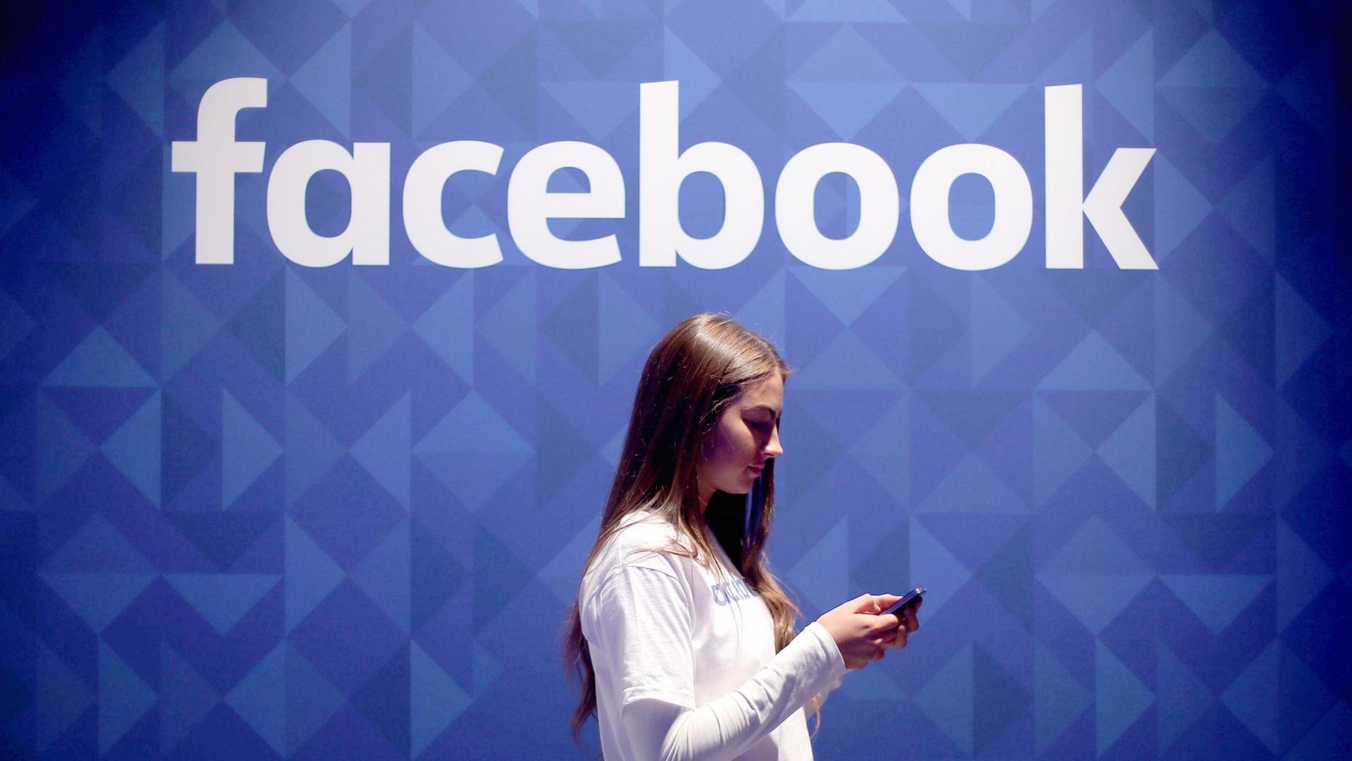 Eine Frau schaut auf ihr Smartphone, während sie an einer blauen Wand mit Facebook-Logo vorbei läuft. Eine Frau schaut auf ihr Smartphone, während sie an einer blauen Wand mit Facebook-Logo vorbei läuft.