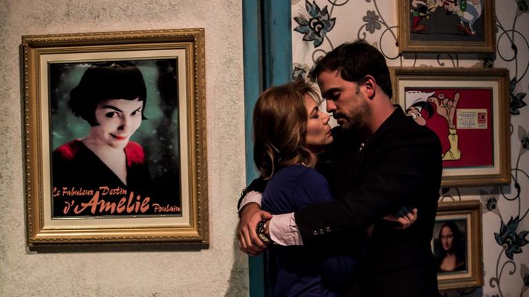 Auf einer Theaterbühne stehen eine Frau und ein Mann und umarmen sich. An der Wand dahinter hängt in einem Bilderrahmen das Filmplakat von "Die fabelhafte Welt der Amelie" Auf einer Theaterbühne stehen eine Frau und ein Mann und umarmen sich. An der Wand dahinter hängt in einem Bilderrahmen das Filmplakat von "Die fabelhafte Welt der Amelie"