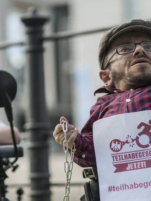 Der Aktivist Raul Krauthausen bei einer Protestaktion gegen das Bundesteilhabegesetz: Er sitzt in einem Rollstuhl, hält einen Zettel mit der Aufschrift "Teilhabegesetz jetzt" in der einen Hand, eine Kette in der anderen.