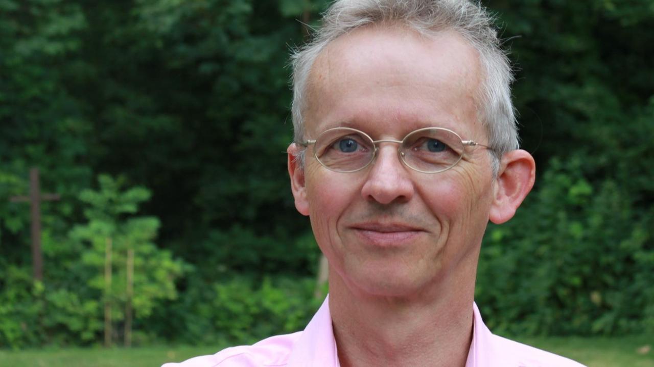 Ein bebrillter Mann mit grauen Haaren und in einem rosafarbenen Hemd steht im Grünen und lächelt in die Kamera. Ein bebrillter Mann mit grauen Haaren und in einem rosafarbenen Hemd steht im Grünen und lächelt in die Kamera.