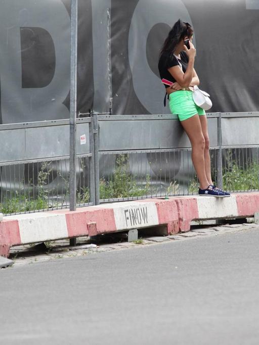 Eine Prostituierte leht sich, mit dem Smartphone in der Hand, an einen Bauzaun in der Kurfürstenstraße, Berlin