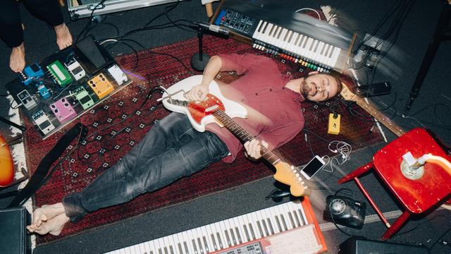 Ein Mann liegt, umringt von Instrumenten, mit seiner E-Gitarre auf dem Rücken auf einem Teppich.