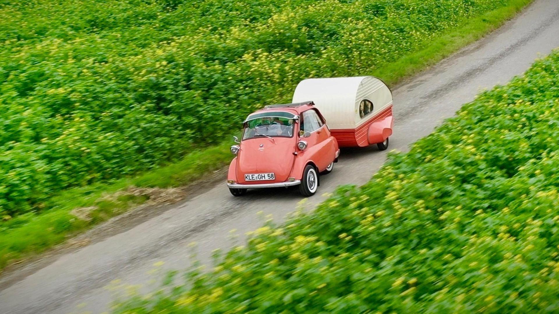 Ein roter BMW Isetta fährt mit dem einem kleinen rot-weißen Wohnwagen entlang grüner Felder.