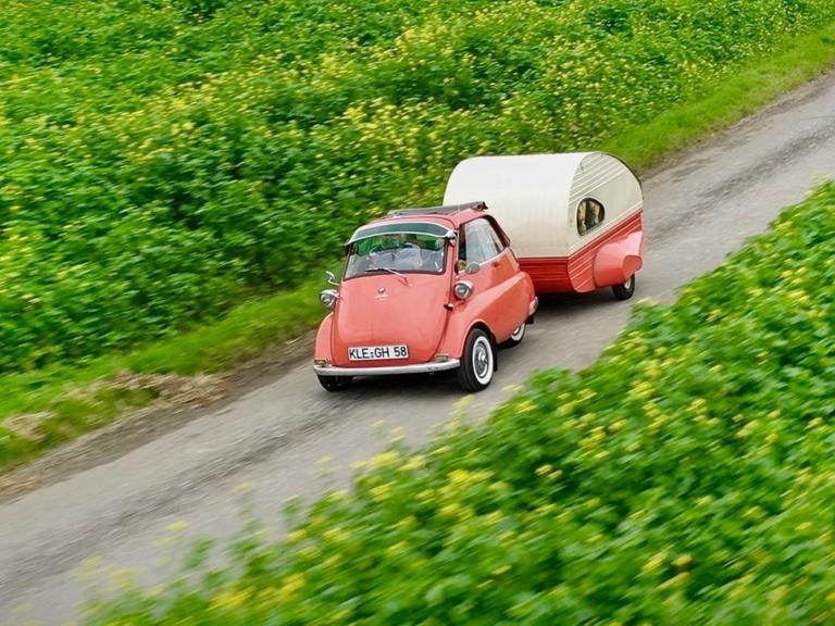 Ein roter BMW Isetta fährt mit dem einem kleinen rot-weißen Wohnwagen entlang grüner Felder.