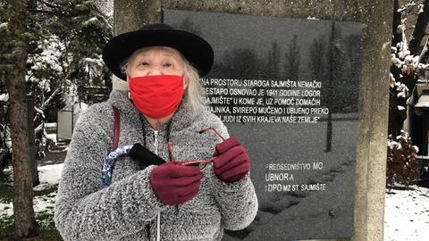 Eine ältere Frau mit Hut, Maske und Krückstock steht vor einer Gedenktafel in Belgrad.