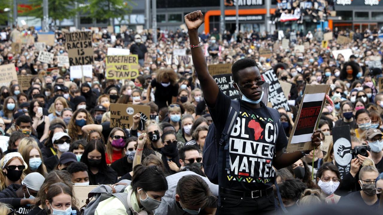Vor einer großen sitzenden Menschenmenge mit Gesichtsmasken steht ein Schwarzer mit erhobener Faust. Auf seinem T-Shirt steht in bunten Buchstaben "Black Lives Matter". Vor einer großen sitzenden Menschenmenge mit Gesichtsmasken steht ein Schwarzer mit erhobener Faust. Auf seinem T-Shirt steht in bunten Buchstaben "Black Lives Matter".