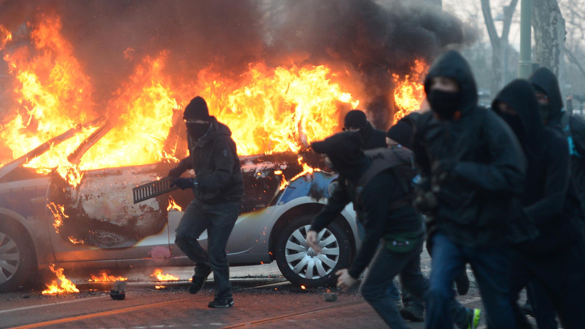 Demonstranten mit schwarzen Kapuzen und Schals, im Hintergrund brennt ein Polizei-Auto Demonstranten mit schwarzen Kapuzen und Schals, im Hintergrund brennt ein Polizei-Auto