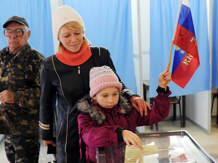 EIne Frau und ein kleines Mädchen in einem Wahllokal. Das Mädchen hält eine Flagge und steckt den Stimmzettel in die Wahlurne.