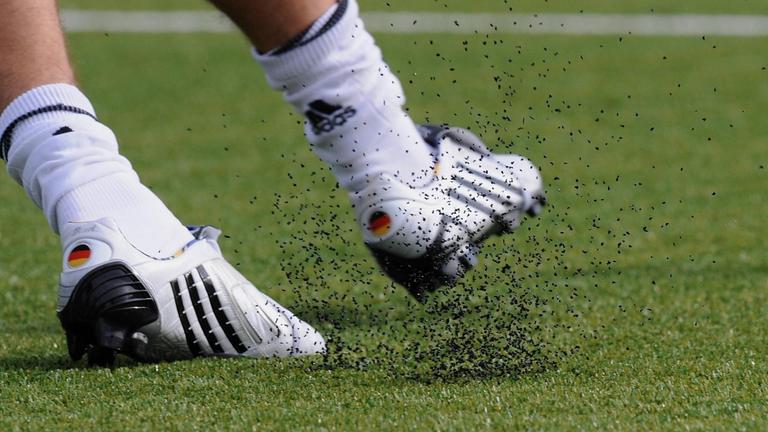 Fußballschuhe mit den deutschen Landesfarben tritt gegen den Ball, dabei löst sich Granulat aus den Kunstrasenhalmen