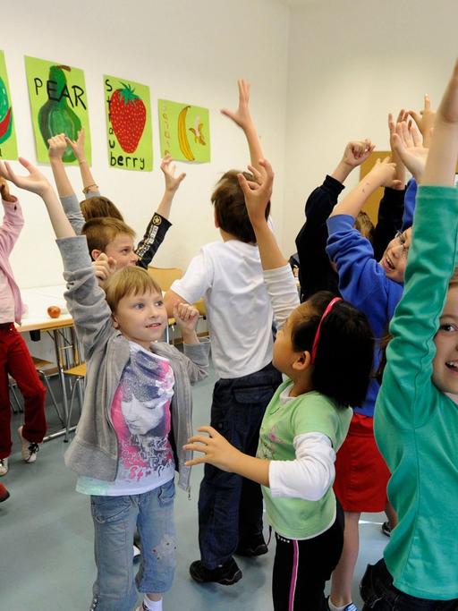 Kinder stehen in einem Klassenraum und heben ihre Arme, an den Wänden bunte Bilder.