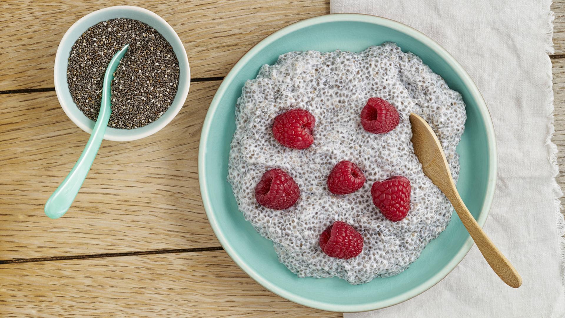 Eine Müslischale mit Chia-Pudding aus Chia-Samen mit Mandelmilch und Himbeeren, daneben eine Schale mit Chia-Samen. Eine Müslischale mit Chia-Pudding aus Chia-Samen mit Mandelmilch und Himbeeren, daneben eine Schale mit Chia-Samen.