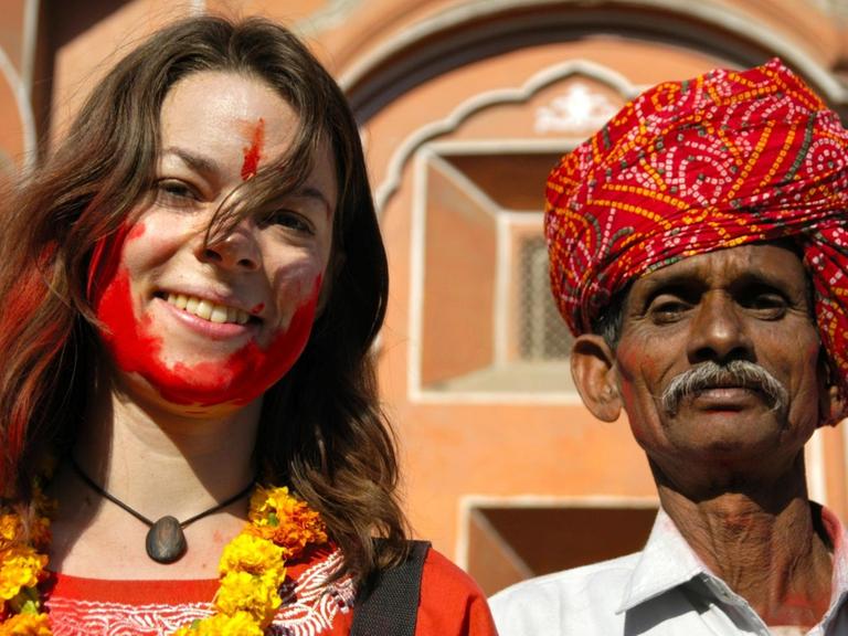 Junge Touristin mit roter Farbe im Gesicht neben einem Rajputen mit rotem Turban vor dem Palast der Winde in Jaipur in Indien.