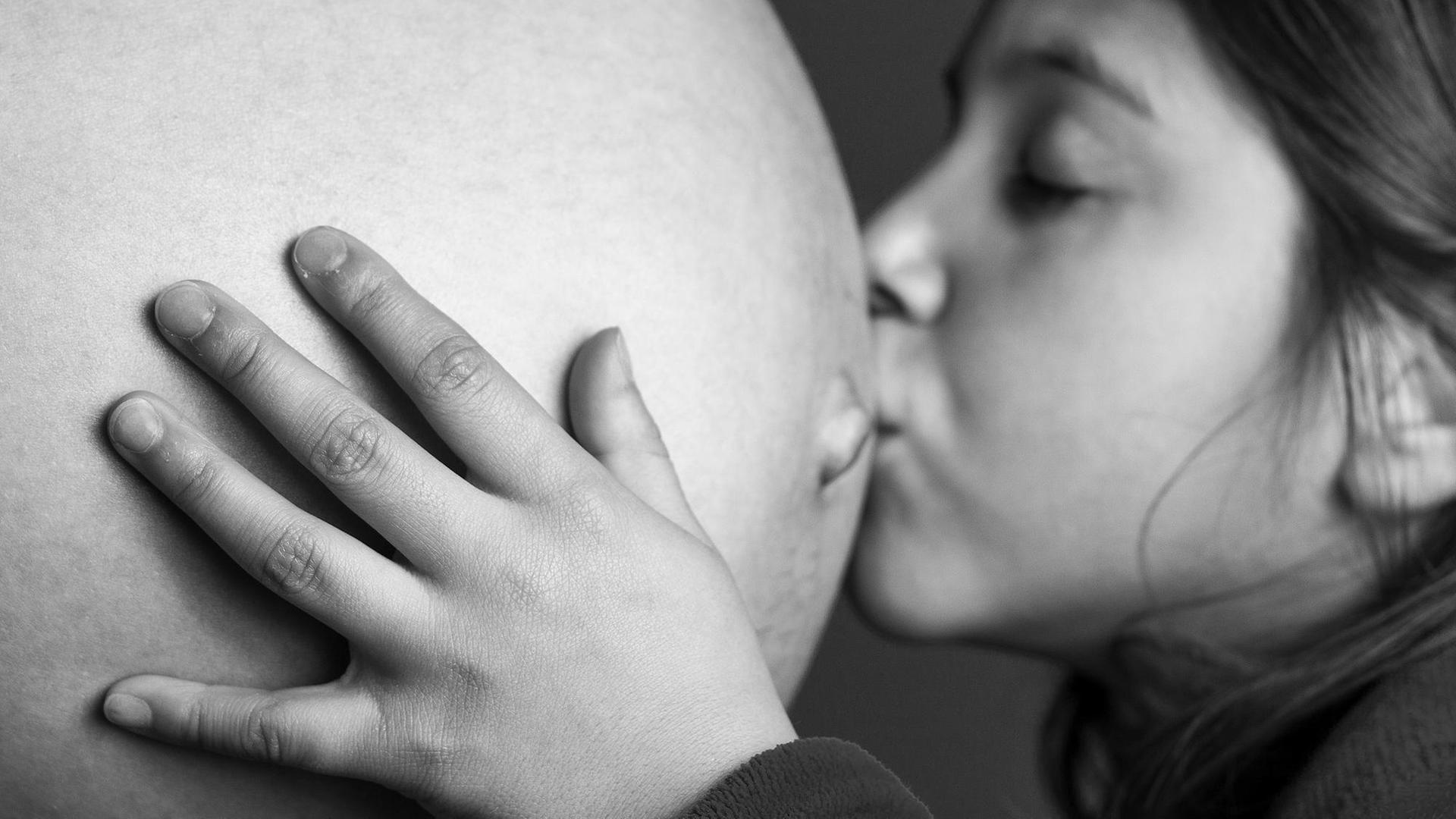 Ein Geschwisterkind küsst den schwangeren Bauch der Mutter und hält ihn liebevoll mit den Händen.