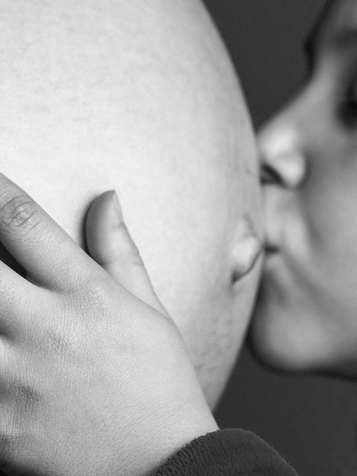 Ein Geschwisterkind küsst den schwangeren Bauch der Mutter und hält ihn liebevoll mit den Händen.