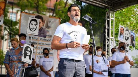 Cetin Gültekin spricht auf einer Freiluftbühne im Sonnenschein in ein Mikrofon. Auf seinen T-Shirt steht der Name seines ermordeten Bruders Gökhan Gültekin sowie eine Zeichnung dessen Gesichts.