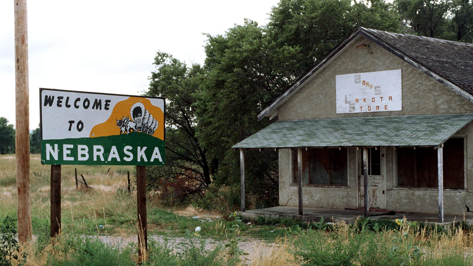 "Welcome to Nebraska" (Willkommen in Nebraska) steht links auf einem Schild an der Staatsgrenze von South Dakota und Nebraska. Rechts ein heruntergekommener, verlassener Laden. Aufgenommen im August 1995 bei Pine Ridge/South Dakota.
