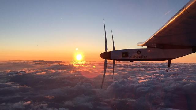 Eine Tragfläche von Solar Impulse 2 mit Rotor über den Wolken vor der untergehenden roten Sonne.