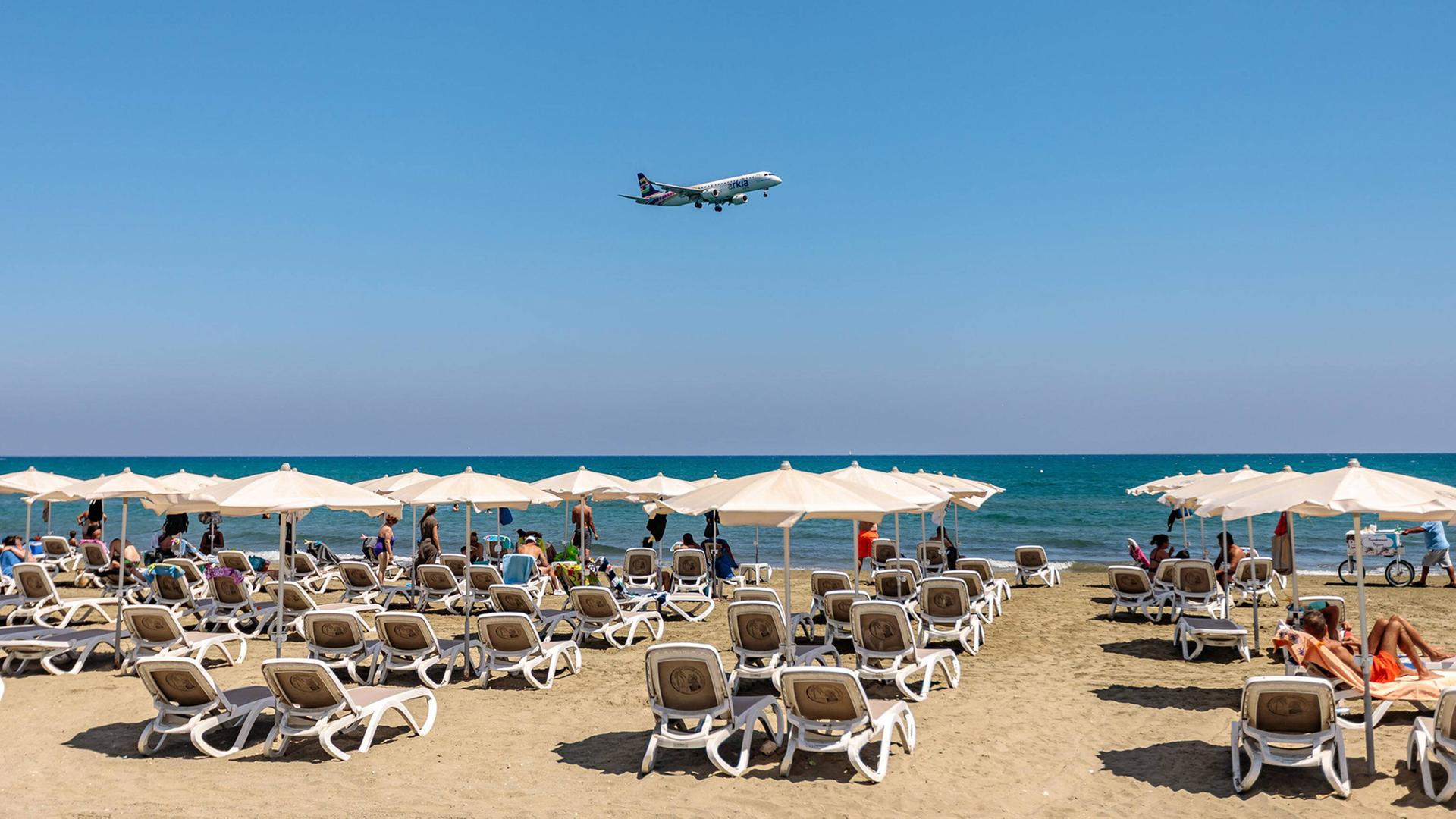 Ein Flugzeug fliegt bei strahlendem Sonnenschein tief über einem Strand mit Reihen voller Liegen und Touristen. Ein Flugzeug fliegt bei strahlendem Sonnenschein tief über einem Strand mit Reihen voller Liegen und Touristen.