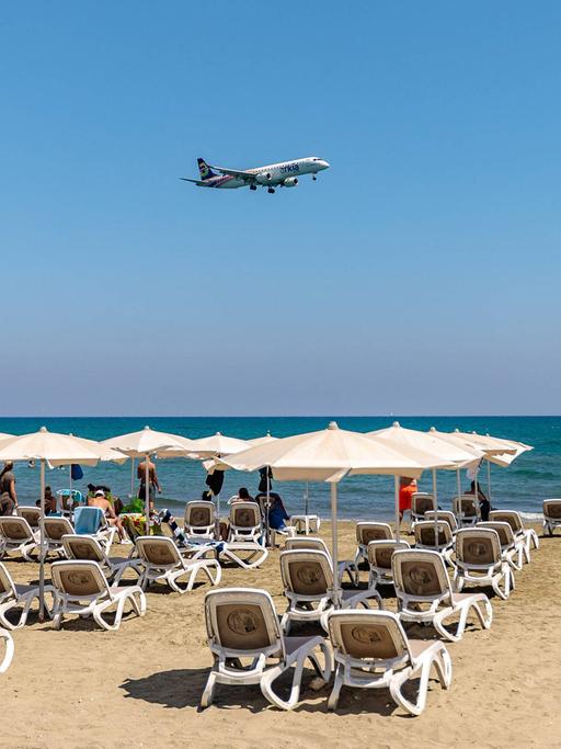 Ein Flugzeug fliegt bei strahlendem Sonnenschein tief über einem Strand mit Reihen voller Liegen und Touristen auf Zypern.
