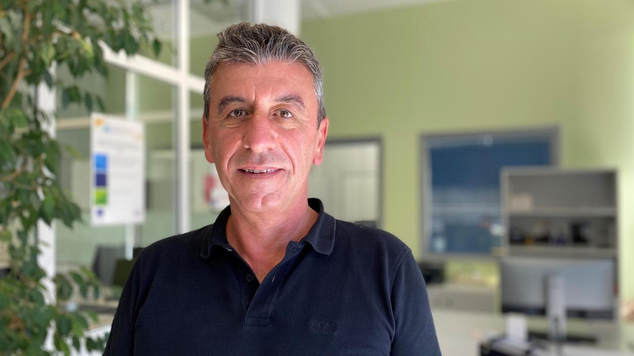 Ein älterer Mann im dunkelblauem T-Shirt und angegrautem Haar steht in einem Büroraum. Ein älterer Mann im dunkelblauem T-Shirt und angegrautem Haar steht in einem Büroraum.