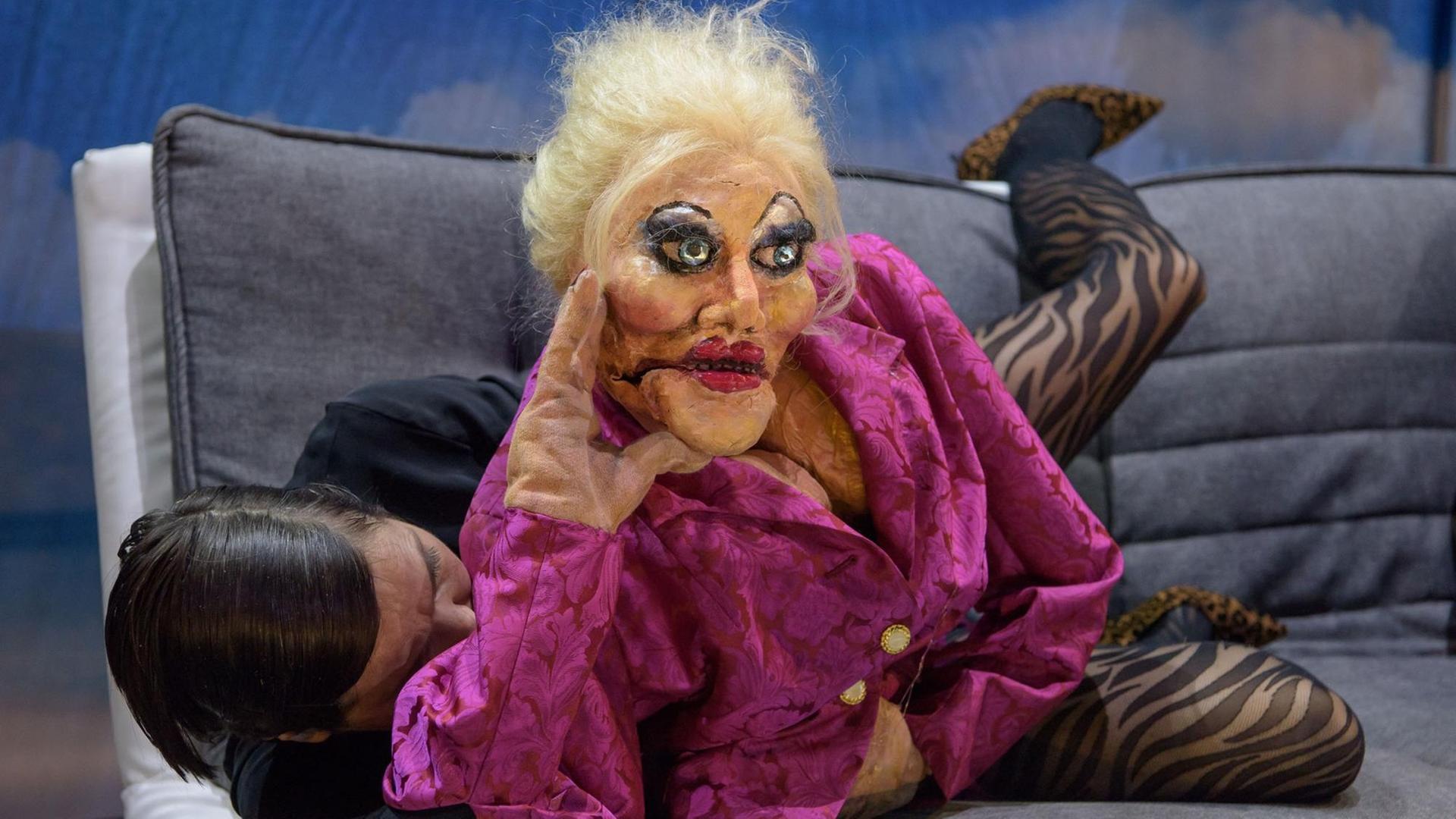 Eine lebensgroße Puppe in einer roten Bluse, aus dem Stück Volksvernichtung , liegt auf einem Sofa und wird von einer Schauspielerin bespielt. Eine lebensgroße Puppe in einer roten Bluse, aus dem Stück Volksvernichtung , liegt auf einem Sofa und wird von einer Schauspielerin bespielt.