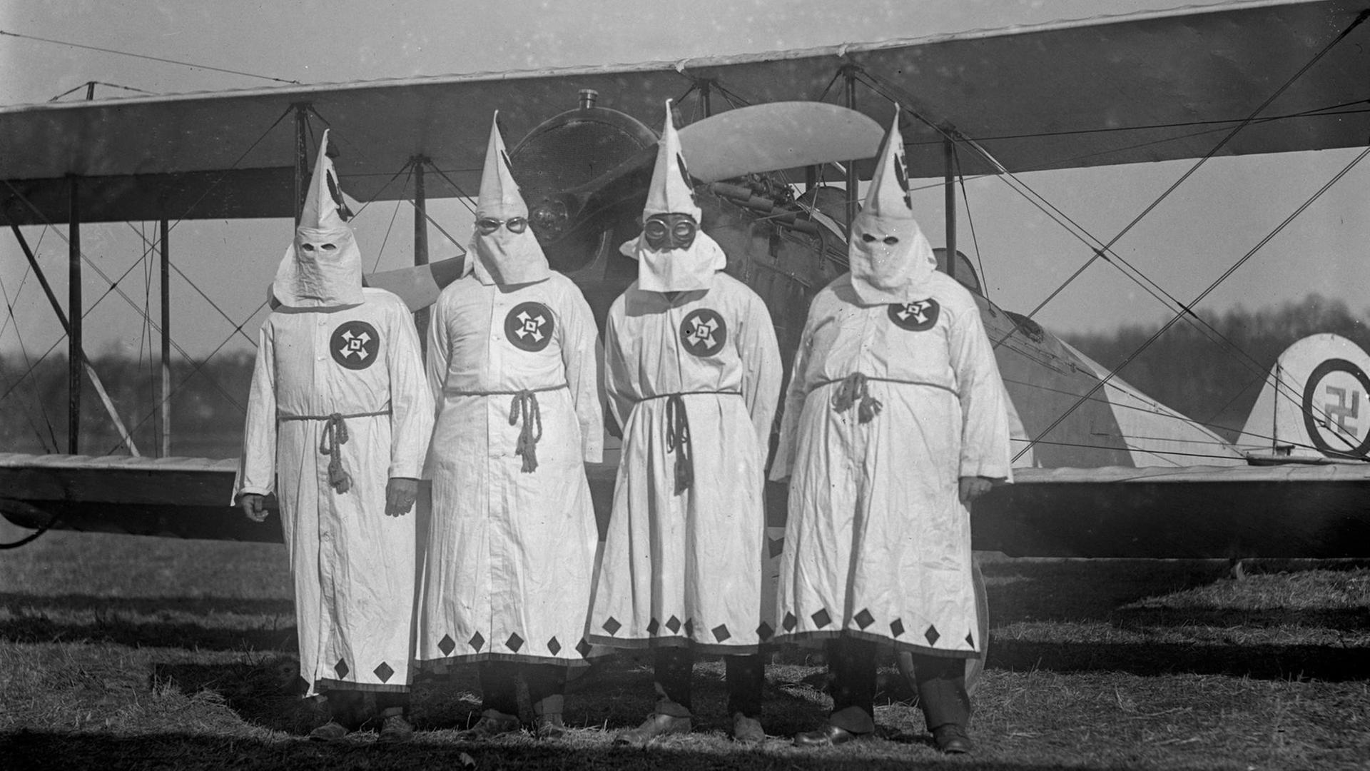 Vier Männer in weißen Kutten mit spitzzulaufenden Hüten stehen vor einer alten Propellermaschine. Das Bild ist schwarz-weiß.