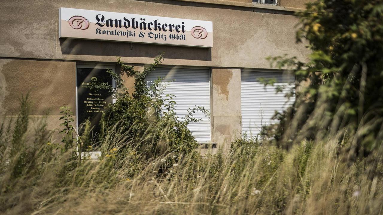 Eine aufgegebene Bäckerei in Klitten. Das Schild des Geschäfts ist noch zu sehen, aber die Rolläden sind herabgelassen und der Zugang zum Gebäude ist von Unkraut überwuchert.