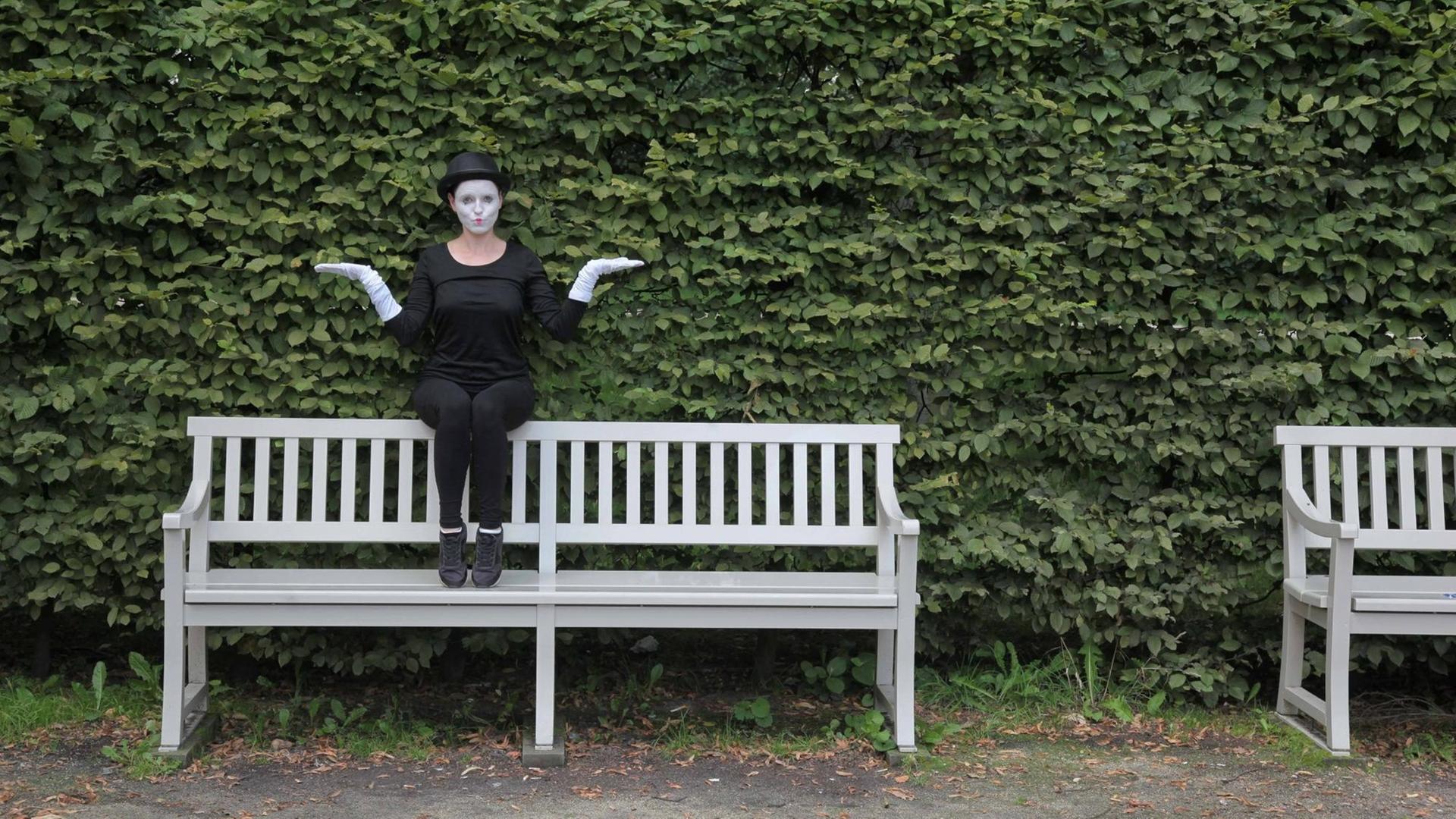 Eine Frau sitzt in Verkleidung einer Pantomime auf der Rückenlehne auf der Parkbank und macht ein witzige Geste. Im Hintergrund ist ein grüner Busch.