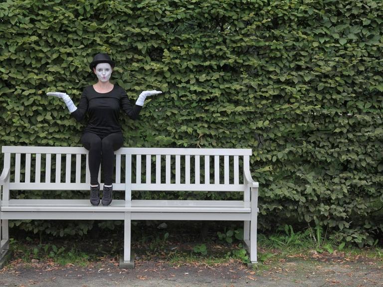 Eine Frau sitzt in Verkleidung einer Pantomime auf der Rückenlehne auf der Parkbank und macht ein witzige Geste. Im Hintergrund ist ein grüner Busch.