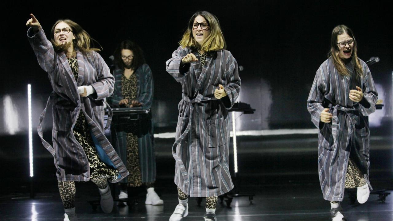 Szene aus dem Theaterstück "Und sicher ist mir die Welt verschwunden". Drei junge Frauen, in Bademänteln und mit Brille, tanzen und singen auf der Bühne.