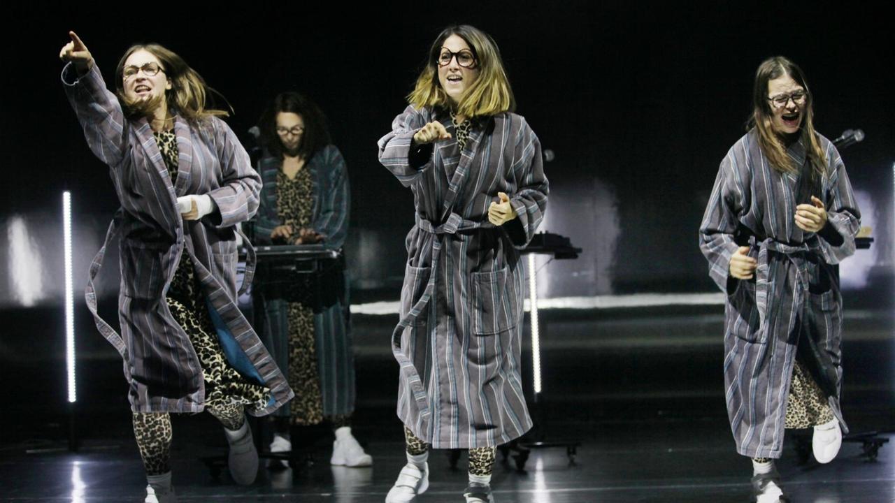 Szene aus dem Theaterstück "Und sicher ist mir die Welt verschwunden". Drei junge Frauen, in Bademänteln und mit Brille, tanzen und singen auf der Bühne. Szene aus dem Theaterstück "Und sicher ist mir die Welt verschwunden". Drei junge Frauen, in Bademänteln und mit Brille, tanzen und singen auf der Bühne.