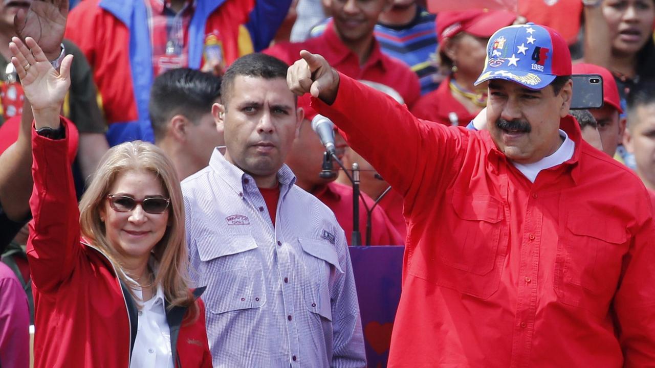 Maduro im roten Hemd und blauer Baseball-Mütze zeigt mit dem Finger nach vorne, links daneben seine blonde Frau mit Sonnenbrille, sie winkt. Dahinter unscharf rotgekleidete Anhänger Maduros.