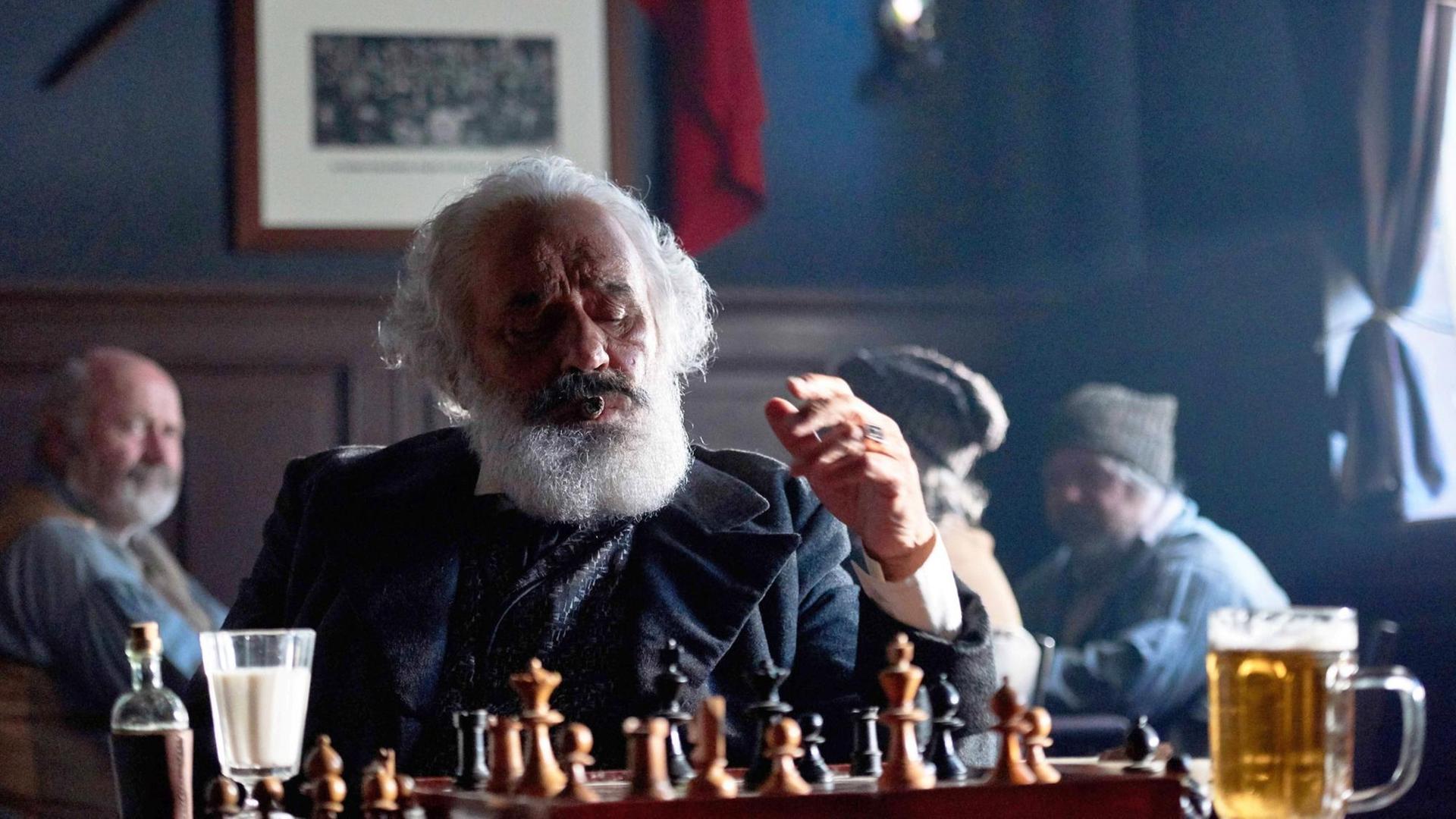 Mario Adorf, in der Rolle des Karl Marx, sitzt mit einem Schachbertt und einem Glas Alkohol am Tisch. Mario Adorf, in der Rolle des Karl Marx, sitzt mit einem Schachbertt und einem Glas Alkohol am Tisch.