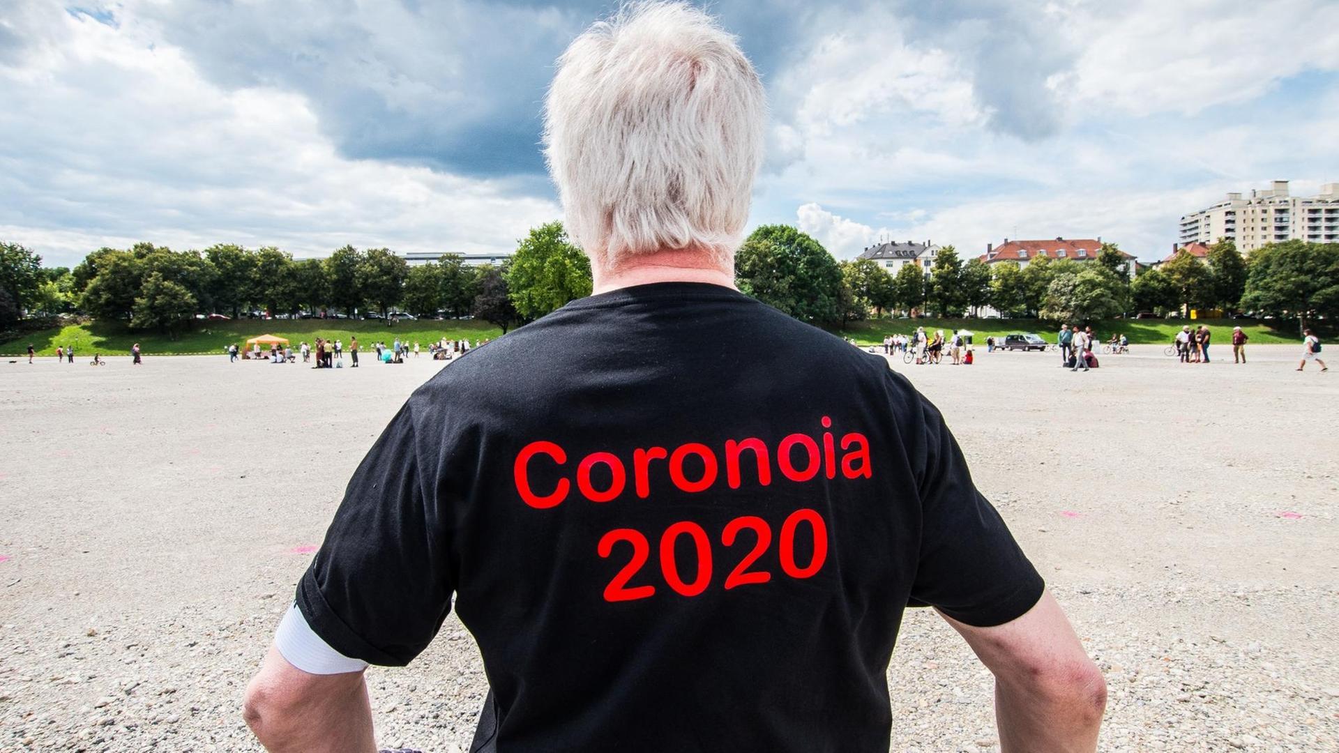 Man sieht einen Mann von hinten, der ein schwarzes T-Shirt mit dem roten Aufdruck "Coronoia 2020" trägt Man sieht einen Mann von hinten, der ein schwarzes T-Shirt mit dem roten Aufdruck "Coronoia 2020" trägt