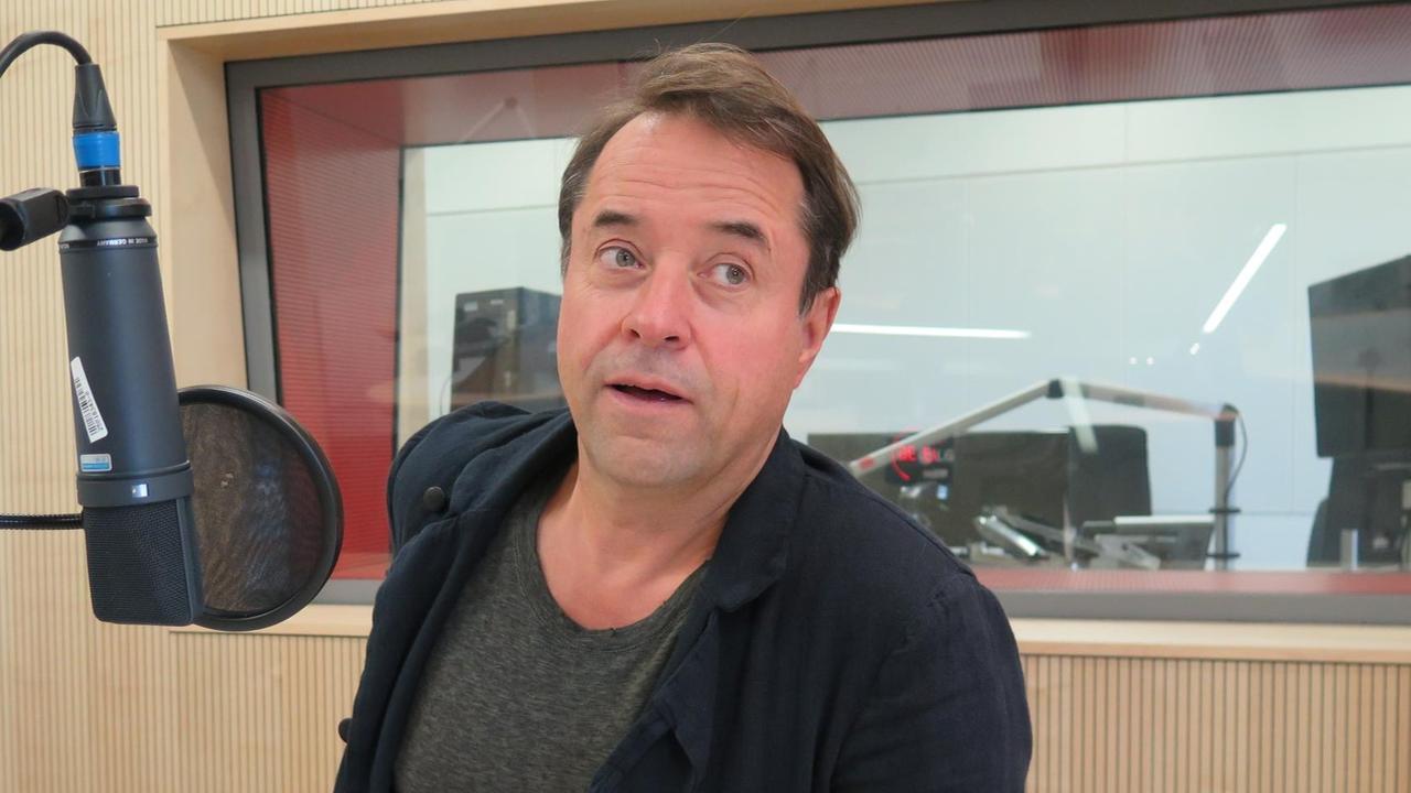 Mann in schwarzem Jackett mit kurzen schwarzen Haaren in Tonstudio hinter einem Mikro, nach oben schauend Mann in schwarzem Jackett mit kurzen schwarzen Haaren in Tonstudio hinter einem Mikro, nach oben schauend