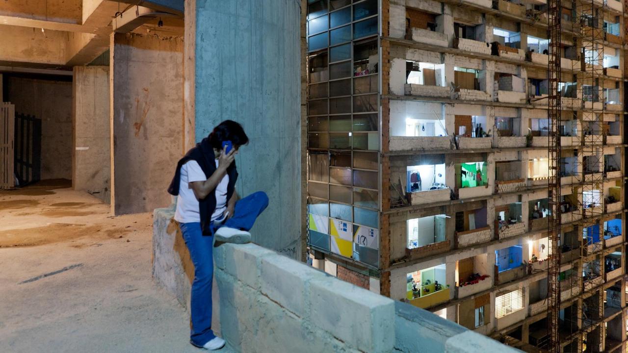 Eine Person sitzt telefonierend auf einer Mauer vor dem Torre David