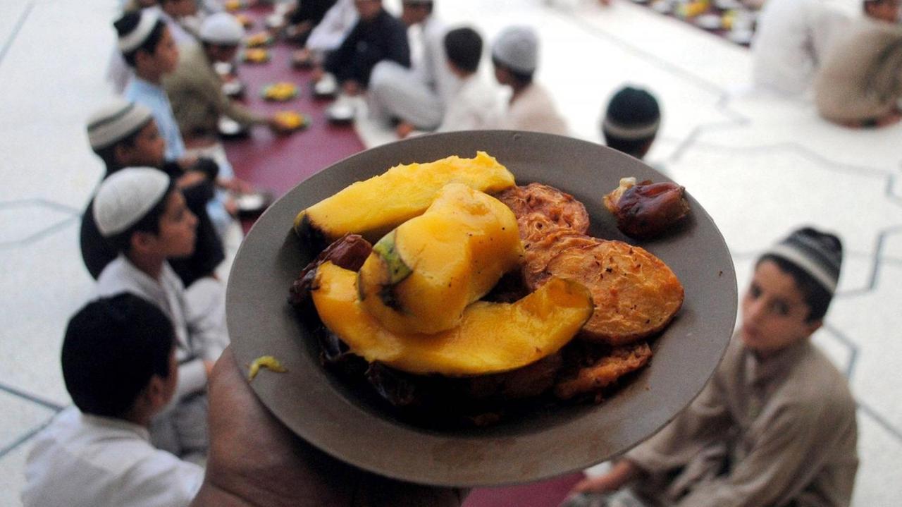 Ein Teller mit Essen ist im Vordergrund. Im Hintergrund sitzen eine Reihe Männer muslimischen Glaubens, die das Fastenbrechen nach Ramadan zelibrieren.