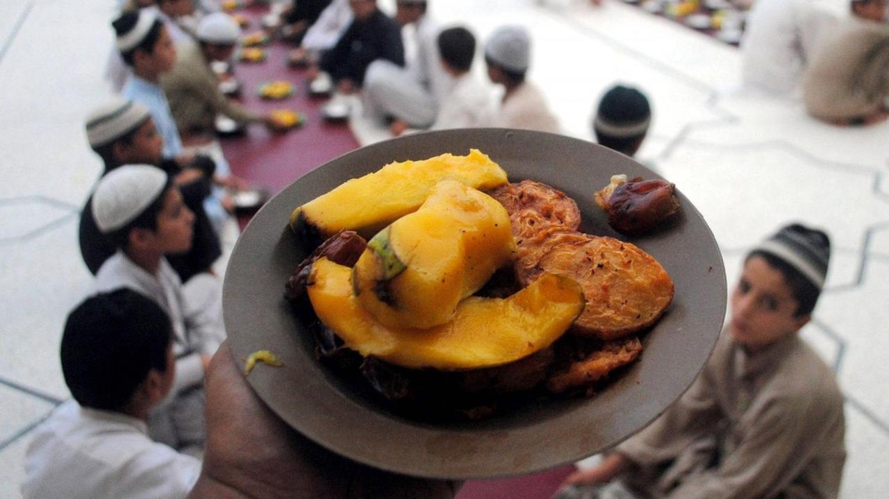 Ein Teller mit Essen ist im Vordergrund. Im Hintergrund sitzen eine Reihe Männer muslimischen Glaubens, die das Fastenbrechen nach Ramadan zelibrieren. Ein Teller mit Essen ist im Vordergrund. Im Hintergrund sitzen eine Reihe Männer muslimischen Glaubens, die das Fastenbrechen nach Ramadan zelibrieren.