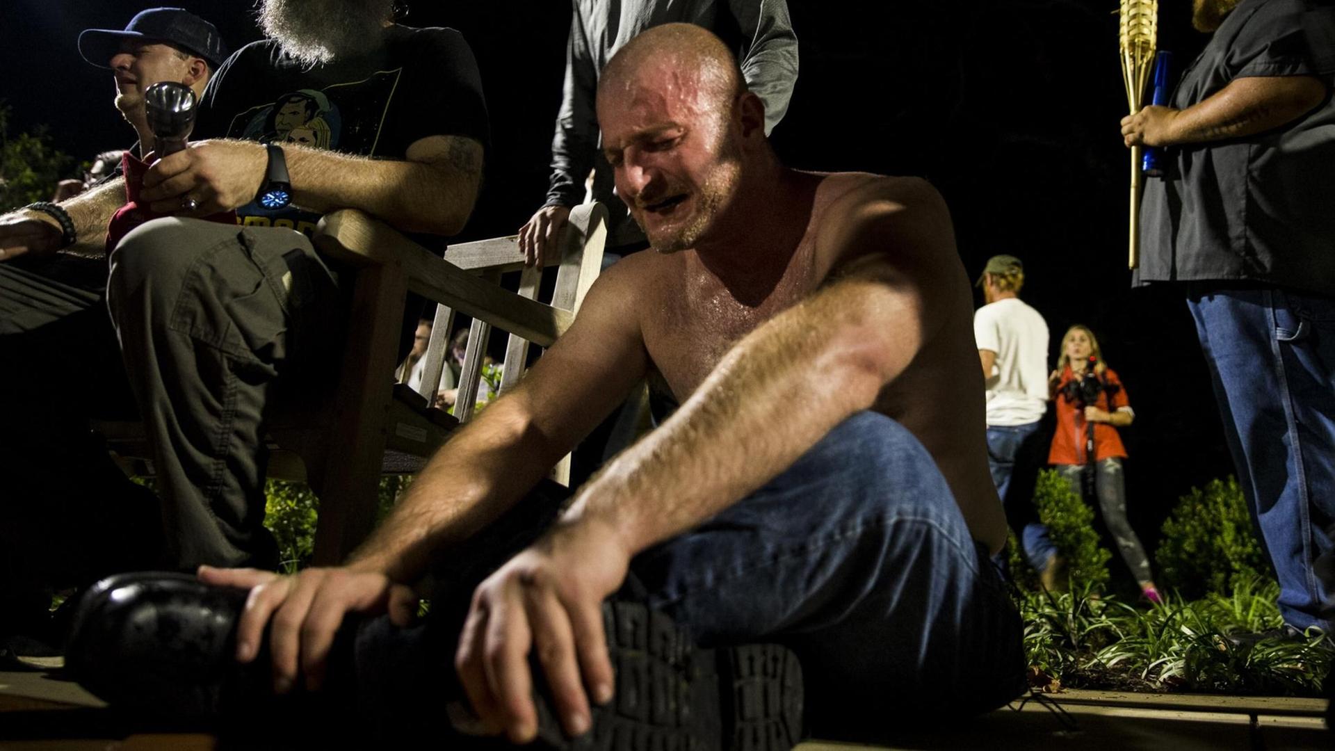 Ein Mann sitzt mit nacktem Oberkörper weinend auf dem Boden, nachdem er von Reizgas getroffen wurde. Um ihn herum stehen ältere Männer mit Fackeln. Ein Mann sitzt mit nacktem Oberkörper weinend auf dem Boden, nachdem er von Reizgas getroffen wurde. Um ihn herum stehen ältere Männer mit Fackeln.