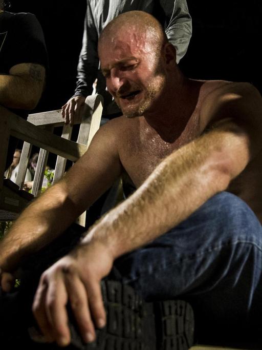 Ein Mann sitzt mit nacktem Oberkörper weinend auf dem Boden, nachdem er von Reizgas getroffen wurde. Um ihn herum stehen ältere Männer mit Fackeln.